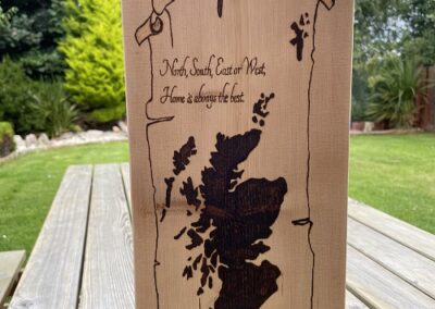 A large piece of wood that has a scroll, the map of Scotland and words that say 'North, South, East or West, Home is always the best.'